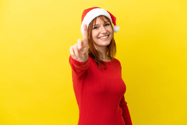 Rudowłosa dziewczyna w świątecznym kapeluszu odizolowany na żółtym tle pokazując i podnosząc palec - Zdjęcie, obraz