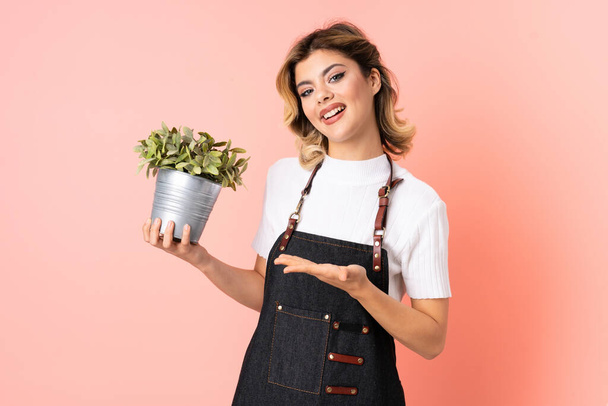 Chica jardinero rusa sosteniendo una planta aislada sobre fondo rosa sosteniendo espacio de copia imaginario en la palma - Foto, Imagen