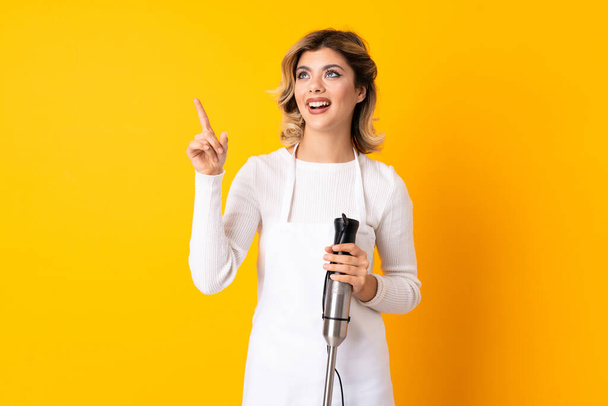 Girl using hand blender isolated on yellow background pointing away - Photo, Image