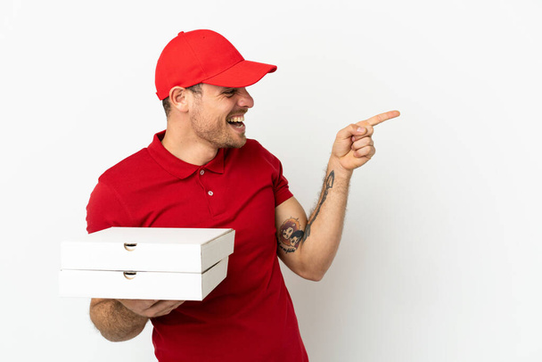 homem de entrega de pizza com uniforme de trabalho pegando caixas de pizza sobre parede branca isolada apontando o dedo para o lado e apresentando um produto - Foto, Imagem