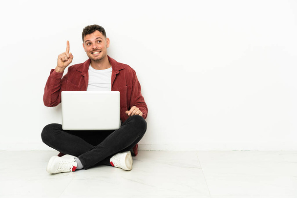 Jonge knappe blanke man sit-in op de vloer met laptop van plan om de oplossing te realiseren terwijl het opheffen van een vinger omhoog - Foto, afbeelding