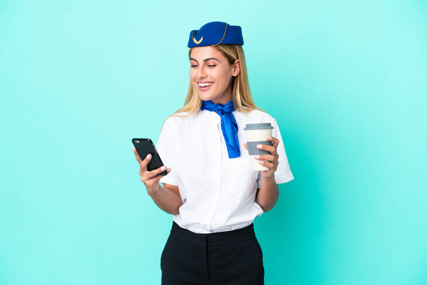 Stewardessa samolotu urugwajska kobieta odizolowana na niebieskim tle trzymająca kawę do zabrania i telefon - Zdjęcie, obraz