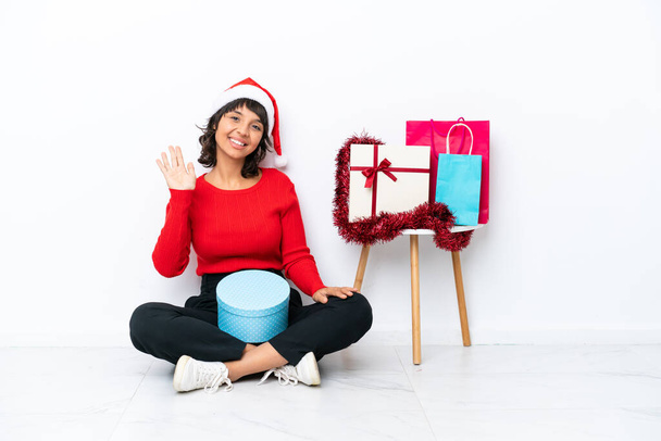 Młoda dziewczyna świętuje Boże Narodzenie siedząc na podłodze odizolowany na białym piekarni salutując z ręki ze szczęśliwym wyrazem twarzy - Zdjęcie, obraz