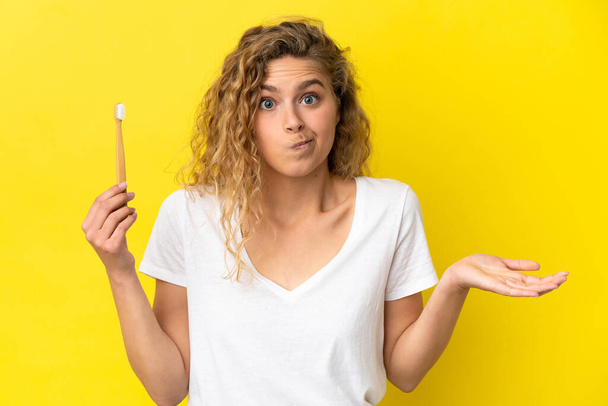 Young caucasian woman holding a brushing teeth isolated on yellow background having doubts while raising hands - Фото, изображение