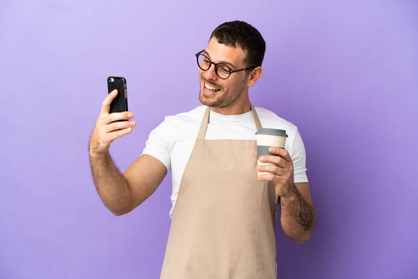 Camarero de restaurante brasileño sobre fondo púrpura aislado sosteniendo café para llevar y un móvil - Foto, Imagen