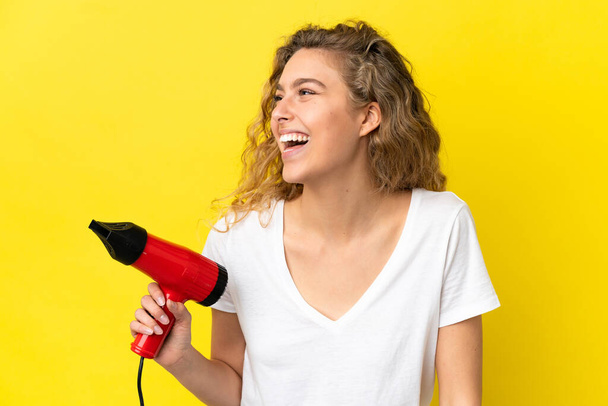 Young blonde woman holding a hairdryer isolated on yellow background laughing in lateral position - Foto, Imagem