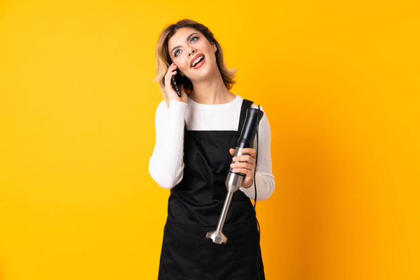 Girl using hand blender isolated on yellow background keeping a conversation with the mobile phone - Photo, Image