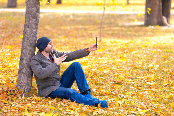 Hombre de mediana edad durante una videollamada en línea en un parque de otoño. Él sentado debajo del árbol en hojas caídas y mira su teléfono inteligente. - Foto, imagen