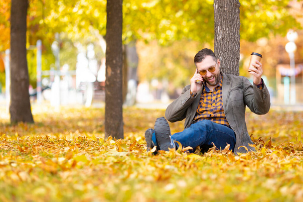 秋の公園でオンラインビデオ通話中に中年の男-彼は落ち葉の上に木の下に座っていると彼のスマートフォンを見て. - 写真・画像