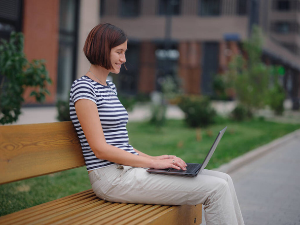 студент, работающий за ноутбуком, садится на скамейку на городской улице. - Фото, изображение