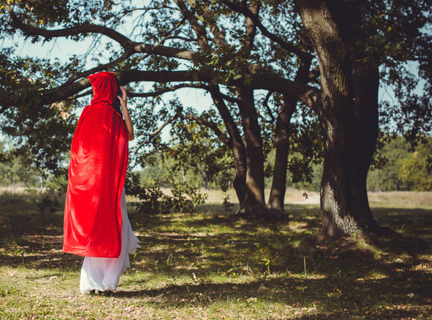 Halloween concept, glamorous costume detail. Young beautiful and mysterious woman in woods, in white Dress and Red Cloak. Little red hood or vampire story - Photo, Image