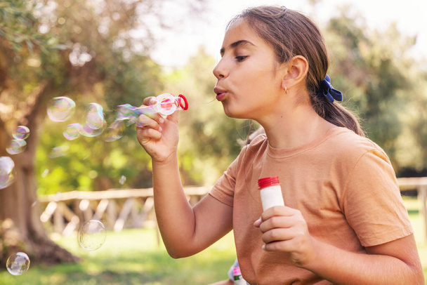 Egy bézs inges kislány szappanbuborékokat fúj a szabadban. Aranyos iskolás lány szórakozik egy parkban nyáron. Gyermek szabadtéri aktivitás koncepció, autentikus gyerekkori pillanat. Póló mockup - Fotó, kép