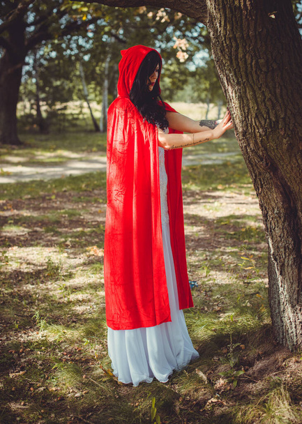 Концепция Хэллоуина, гламурная деталь костюма. Молодая красивая и таинственная женщина в лесу, в белом платье и красном плаще. Красный капюшон или вампирская история - Фото, изображение