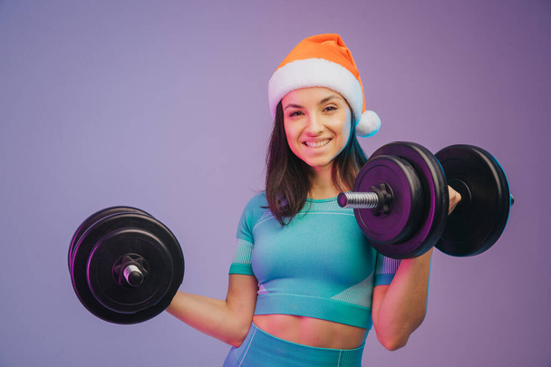 Close up portrait of young fit and sportive woman in Christmas hat, on gradient background. Brunette longhair model. Perfect body ready for summertime. Beauty, resort, sport concept - Foto, afbeelding