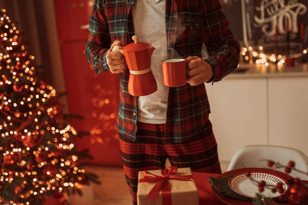 человек в пижаме проведение кофе в красной чашке с гейзером кофеварка в уютной праздничной атмосфере Рождества - Фото, изображение