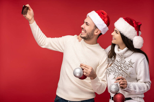 Photo of sweet couple in Christmas hats and with Christmas toys, looks happy. Male and female take a selfie and smile, isolated over red background - Photo, image