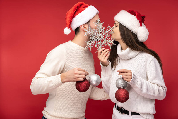 Photo of sweet couple in Christmas hats and with Christmas toys, kiss each other. Male and female are in love look blessed and smile, isolated over red background - Photo, Image