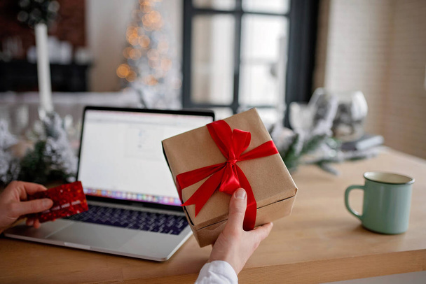 Adult man holds a gift for advent holiday on a laptop background. Makes online purchases. Happy New Year and Merry Christmas - Foto, afbeelding
