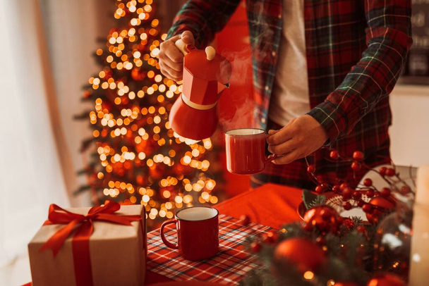 hombre en pijama verter el café en la taza roja de la cafetera géiser en un acogedor ambiente navideño festivo - Foto, imagen
