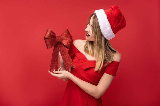 Noel şapkalı ve kırmızı elbiseli çekici bir kadının fotoğrafı Noel ağacı için kırmızı fiyonk tutuyor ve şuna bak. Kırmızı arkaplan üzerinde izole edilmiş - Fotoğraf, Görsel