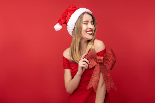 Photo of attractive woman in Christmas hat and red dress, holds red bow for Christmas tree and smile. Isolated over red background - Photo, Image