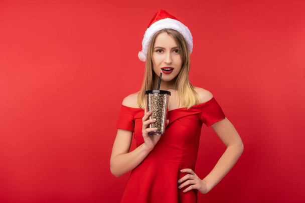 Foto di donna attraente in cappello di Natale e vestito rosso, tiene la tazza di plastica di caffè e posa mano su fianchi. Isolato su sfondo rosso - Foto, immagini