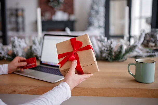 Adult man holds a gift for advent holiday on a laptop background. Makes online purchases. Happy New Year and Merry Christmas - 写真・画像