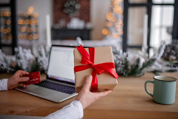 Adult man holds a gift for advent holiday on a laptop background. Makes online purchases. Happy New Year and Merry Christmas - Foto, immagini