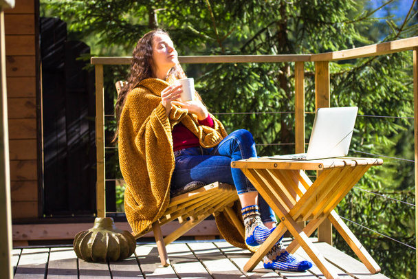 Pretty white girl is sitting relaxing on balcony wrapped in yellow plaid holding cup of tea coffee. Young brunette woman works remotely on laptop, shopping online. Sweet home in a wood at autumn day. - Фото, изображение