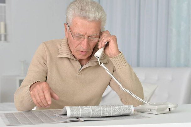 Senior man reading newspaper and talking on phone at home - Foto, Imagen