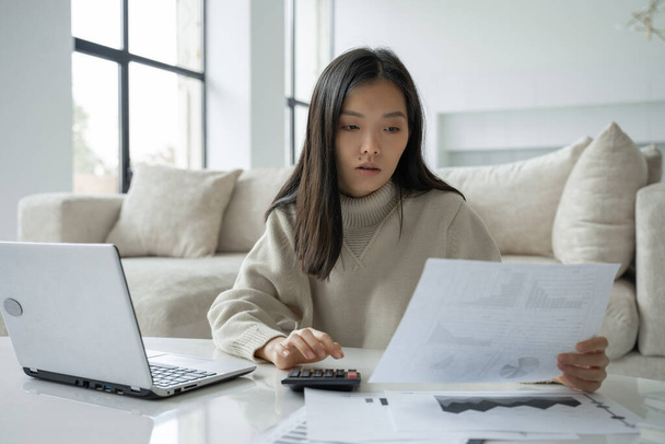 Asian woman with a laptop and a calculator in the living room. A serious lady with the help of a laptop computer calculates finances at home and looks at her bills on paper - Фото, зображення