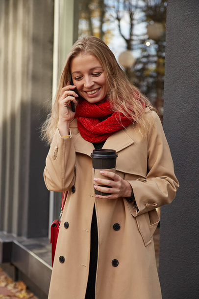 ベージュトレンチコートを着た白人女性がコーヒーを片手にスマートフォンを呼び - 写真・画像