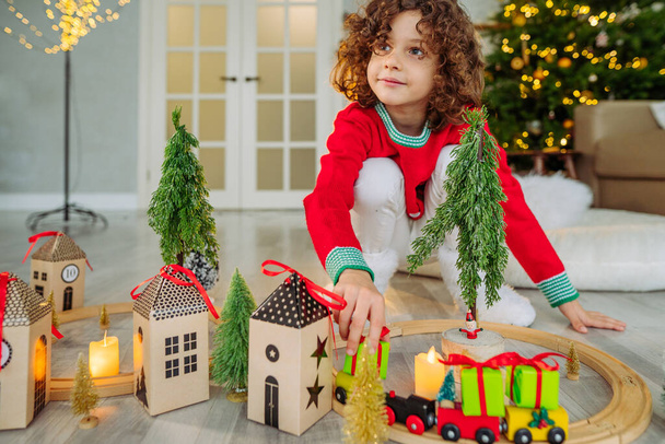 Happy little boy with curly hair playing with wooden railroad at Christmas time at home. - Photo, Image