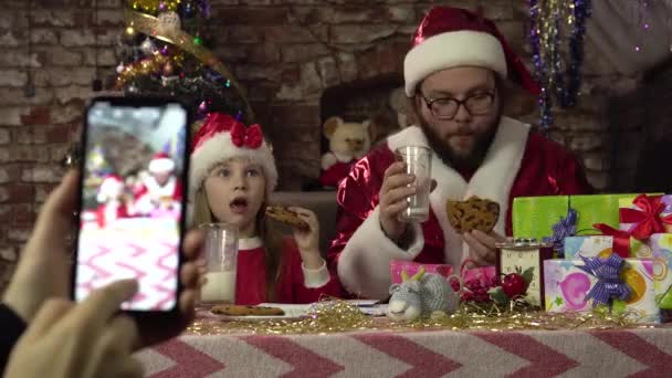 Un hombre vestido de Santa y una niña están bebiendo leche y cookies.Posing para el teléfono inteligente. - Imágenes, Vídeo