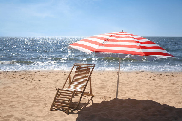 Deck chair near red and white striped beach umbrella on sandy seashore - Valokuva, kuva