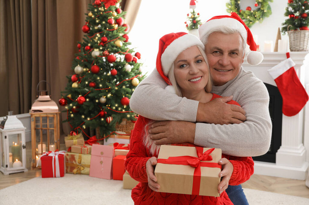 Happy mature couple in Santa hats with gift box at home, space for text. Christmas celebration - Φωτογραφία, εικόνα