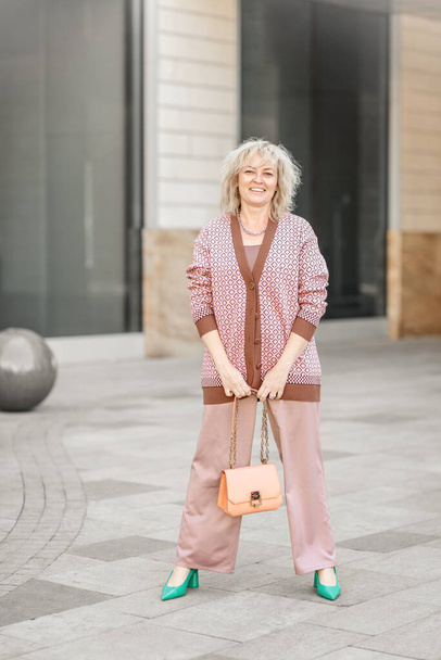 Ελκυστική ενήλικη ξανθιά ευρωπαϊκής εμφάνισης με παντελόνι και τσάντα στα χέρια της στους δρόμους της πόλης - Φωτογραφία, εικόνα