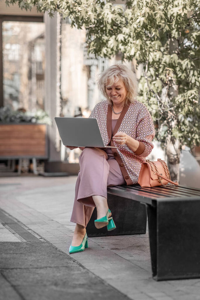 Attractive adult blonde of European appearance sitting on a bench in the city leads a video conference online with a laptop in her hands - Фото, зображення
