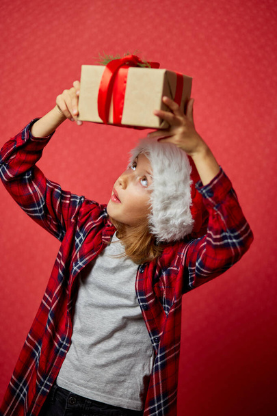 Meglepett és pozitív kislány télapó kalap kezében karácsonyi ajándék és ellenőrizze az ajándék doboz, elszigetelt piros háttér, boldog új évet - Fotó, kép