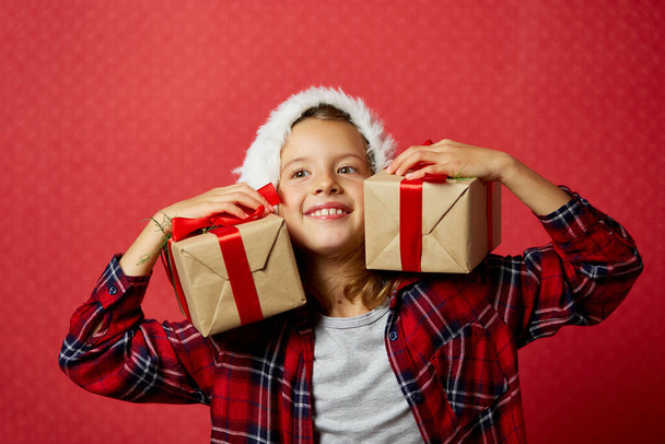 Здивована і позитивна дівчинка в капелюсі Санта тримає два різдвяні подарунки, подарункові коробки, ізольовані на червоному тлі, щасливий Новий рік
 - Фото, зображення
