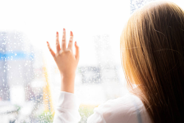 Przygnębiona młoda kobieta w pobliżu okna w domu, zbliżenie, smutna młoda kobieta patrząc przez okno z kroplami deszczu - Zdjęcie, obraz