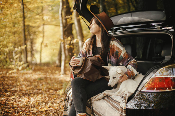 Mulher viajante elegante com mochila sentada com cão bonito no porta-malas do carro em florestas de outono ensolaradas. Jovem hipster fêmea viajando com cão branco pastor suíço. Viagem e viagem de carro com animal de estimação - Foto, Imagem