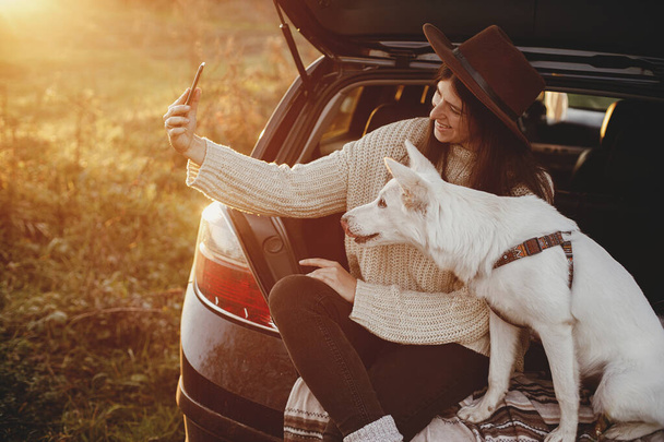 Tyylikäs hipster nainen ottaa selfie valokuva söpö koira auton takakontissa lämpimässä auringonlaskussa. Syksyllä road trip lemmikin kanssa. Nuori nainen hattu ja pusero puhelimitse ja matkustaa makea valkoinen koira - Valokuva, kuva