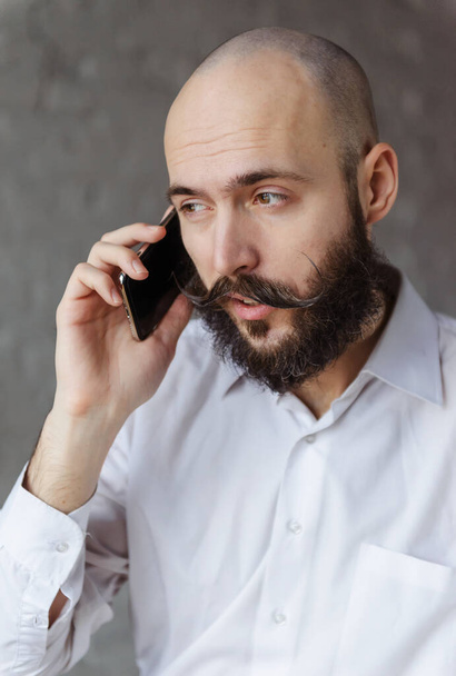 giovane uomo calvo con la barba e baffi in camicia bianca sta parlando al telefono - Foto, immagini
