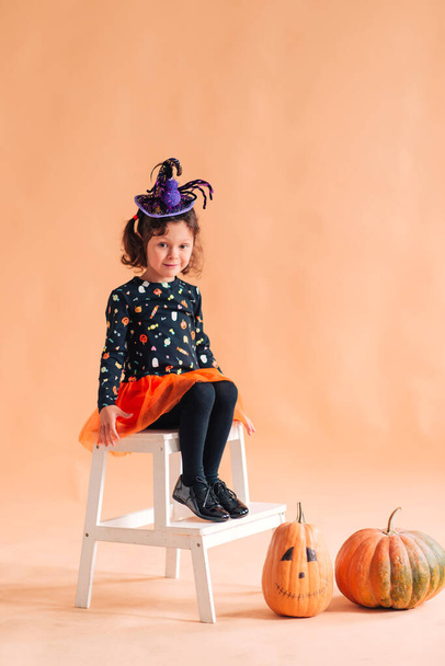 Funny little girl in a witch costume and a hat with a spider for Halloween with a pumpkin Jack on a plain background - Valokuva, kuva