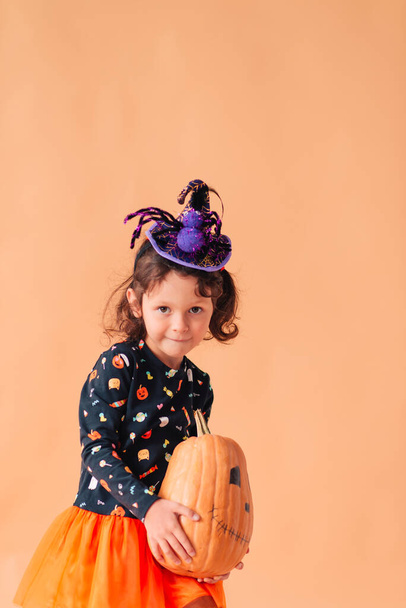 Funny little girl in a witch costume and a hat with a spider for Halloween with a pumpkin Jack on a plain background - Foto, imagen