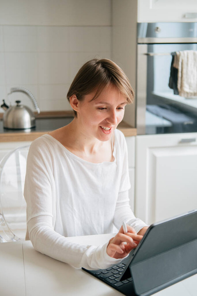 Portrait of young beautiful woman in white shirt using computer, sitting in kitchen in modern apartment. Remote work and freelance concept. - Foto, imagen
