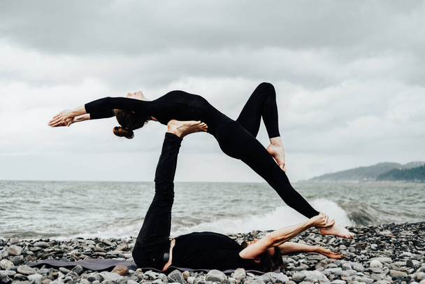 Man and woman in black sport clothes doing acroyoga on a sea beach. Woman lying on male feet. Healthy lifestyle, yoga lovers. - Φωτογραφία, εικόνα
