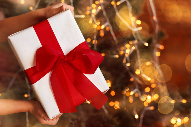 Christmas gift box with a red bow in children's hands on an illumination background - 写真・画像