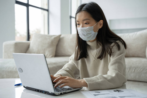 Een attente Aziatische vrouw met een medisch masker met behulp van een laptop werkt in de woonkamer. Studenten online, afstandsonderwijs - Foto, afbeelding